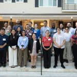Group of newly hired faculty members pose together on the front steps of a campus residence during a welcome gathering, with the building entrance behind them.