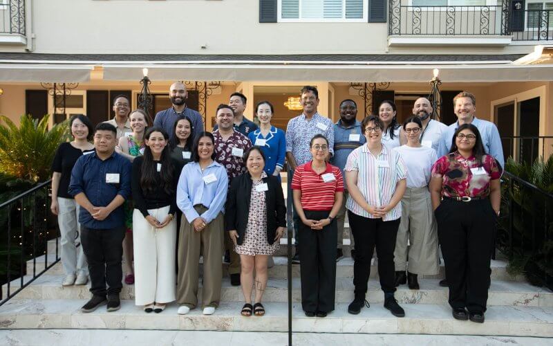 Group of newly hired faculty members pose together on the front steps of a campus residence during a welcome gathering, with the building entrance behind them.