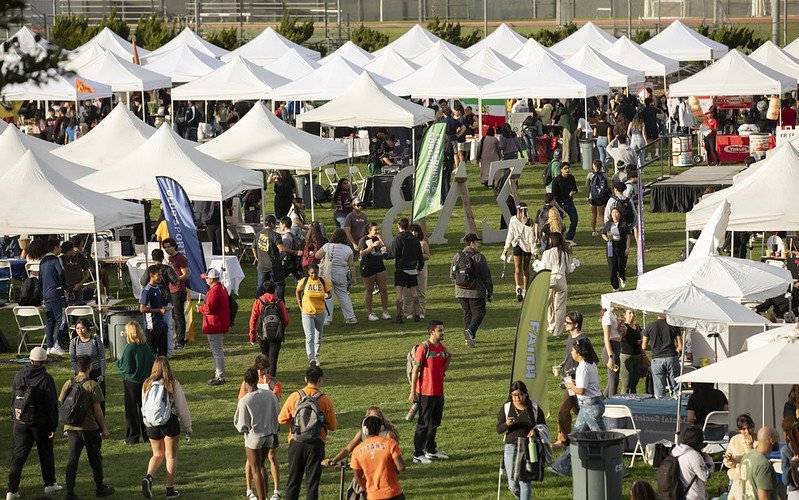 Students walk among rows of white canopy tents at Discoverfest on the CSUF intramural field, where student organizations host tables and displays.