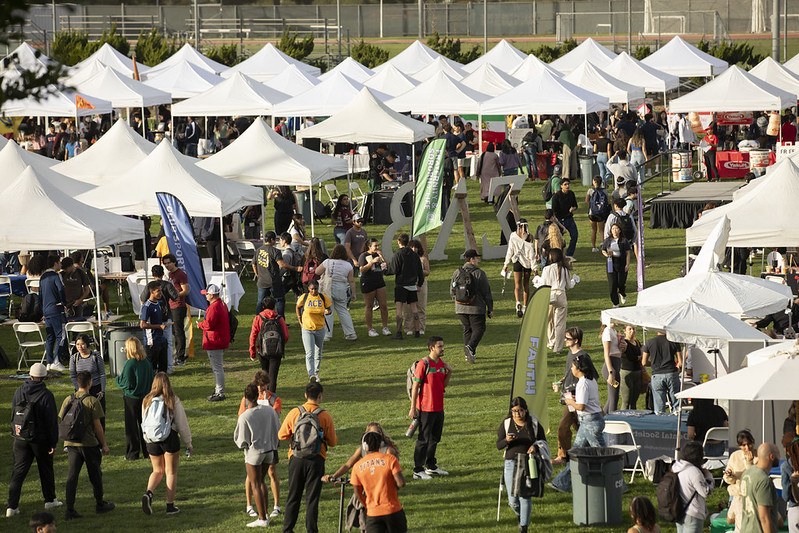 Students walk among rows of white canopy tents at Discoverfest on the CSUF intramural field, where student organizations host tables and displays.