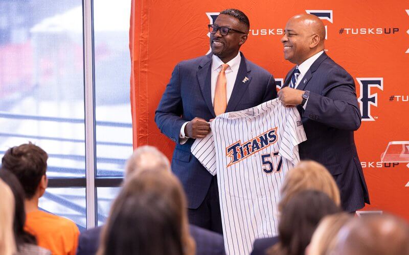 Two men in business attire smile while holding a Titans baseball jersey during a campus announcement, with attendees seated in the foreground and a Cal State Fullerton backdrop behind them.