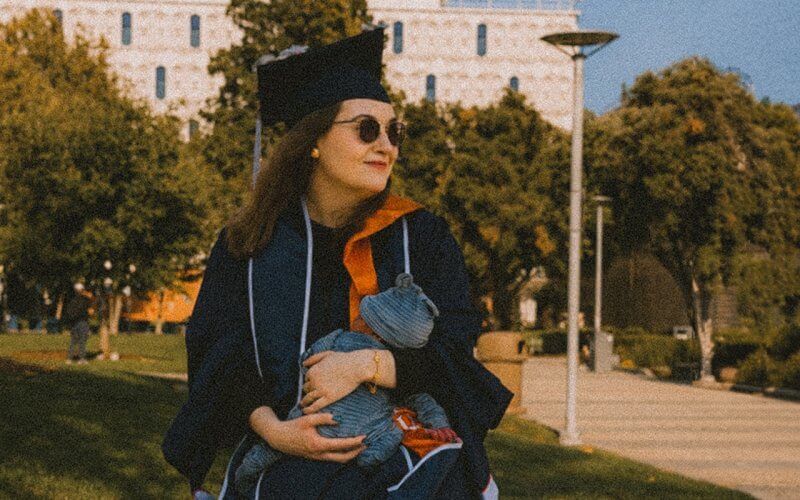 Alina Chupikova holds her newborn child while wearing CSUF graduation regalia.