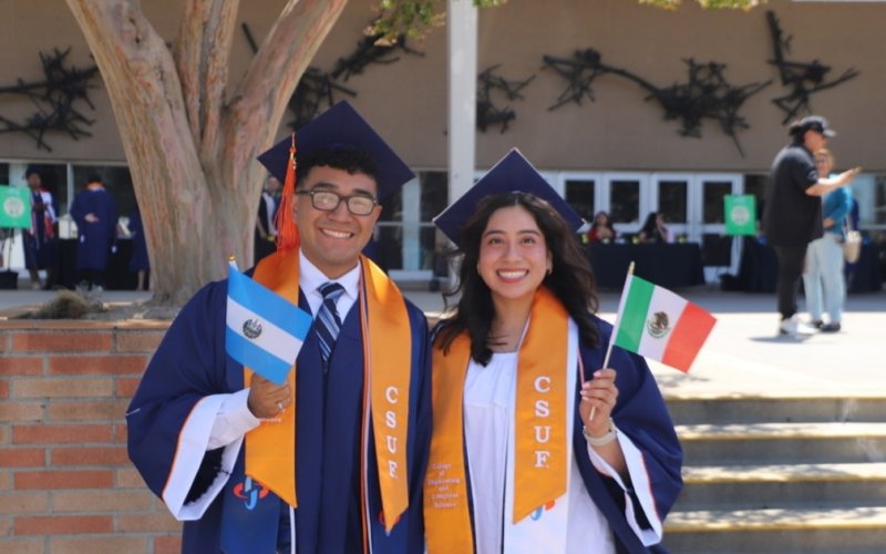 Two students in graduation regalia, one holding a Mexico flag and the other an El Salvador flag, smile at the camera.