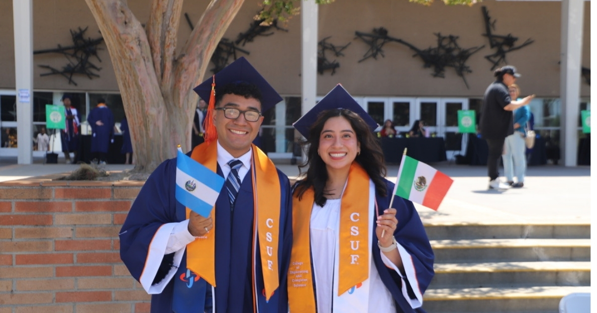 Two students in graduation regalia, one holding a Mexico flag and the other an El Salvador flag, smile at the camera.