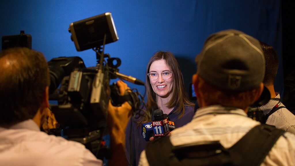 Faculty member speaks with television reporters while cameras and microphones from local news stations surround her during a media interview.