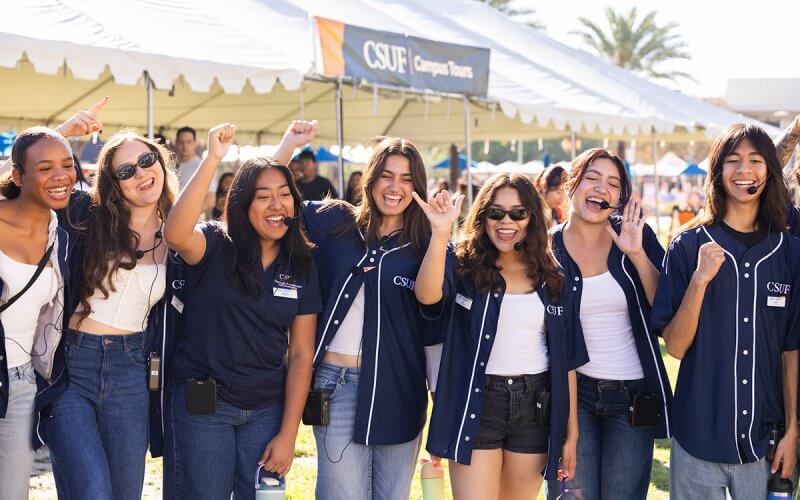 Alt text: Seven smiling Cal State Fullerton student volunteers wearing headsets cheer and raise their hands in front of a CSUF Campus Tours tent at an outdoor event.